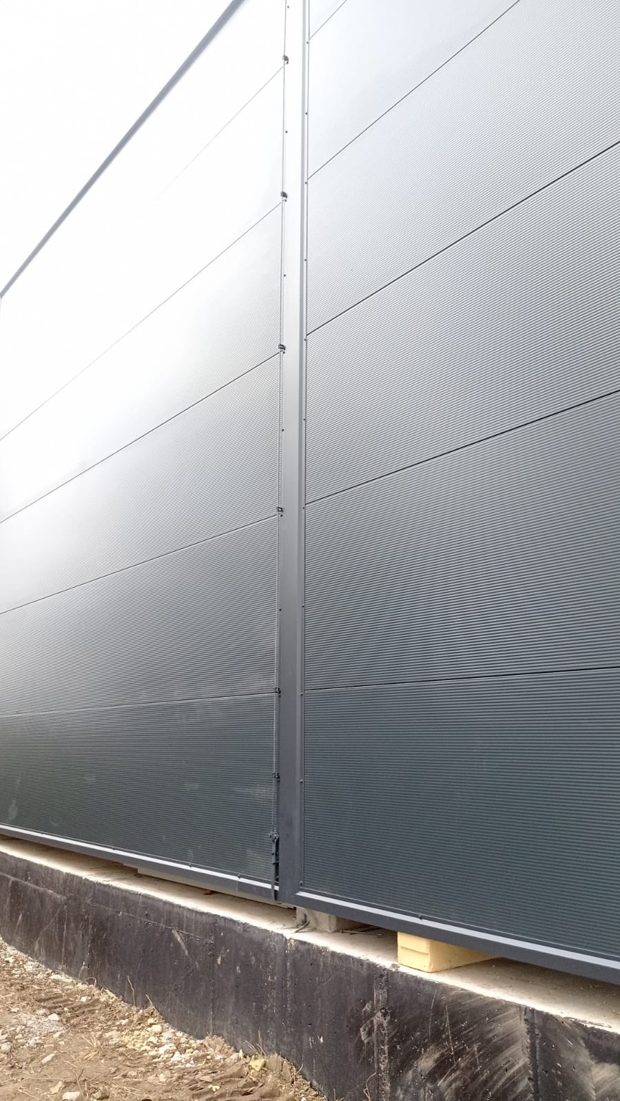Szara, falista blacha trapezowa na elewacji budynku przemysłowego, widoczne łączenia paneli i podmurówka.