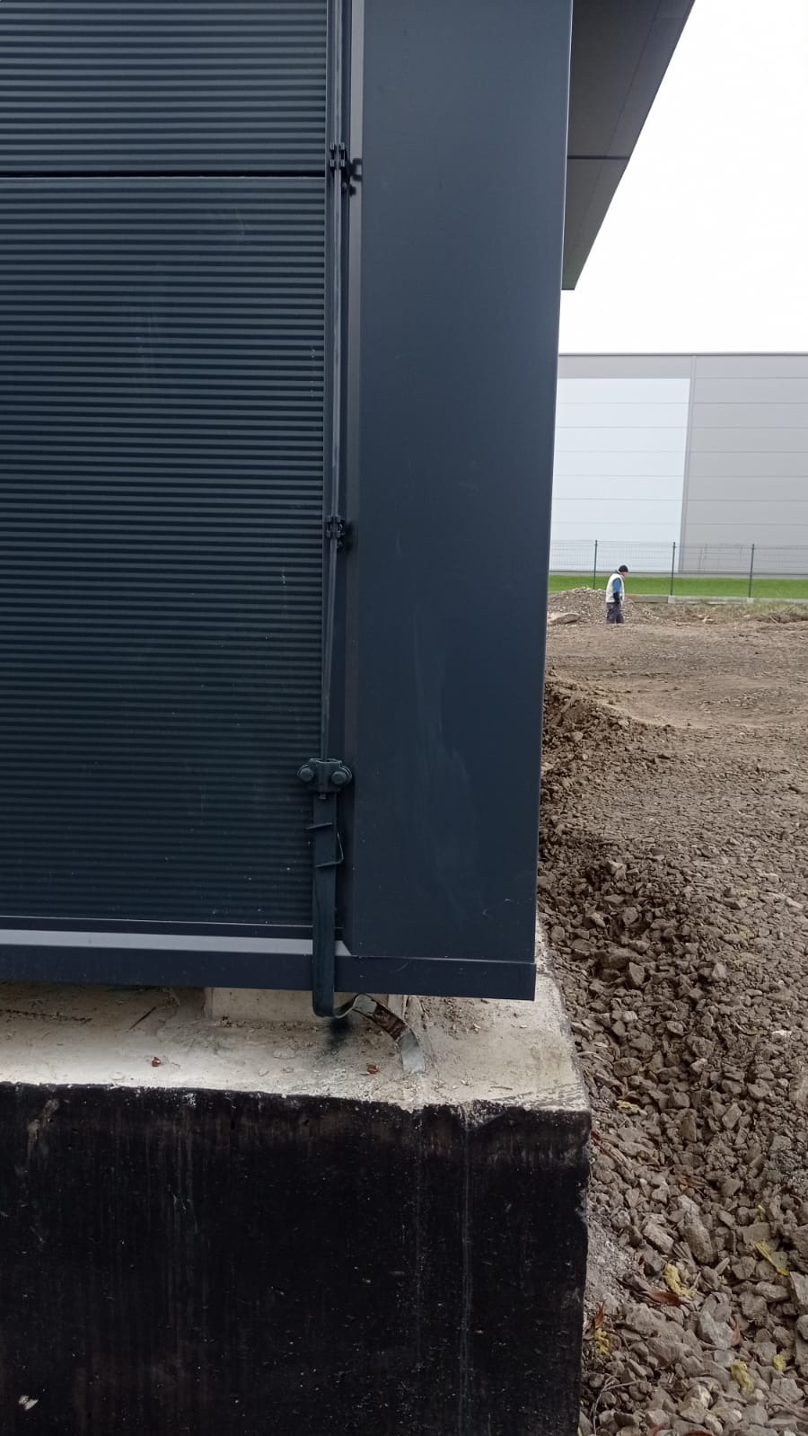 Fragment hali przemysłowej z widocznym systemem odprowadzania ładunków elektrycznych, ziemia wokół w trakcie prac ziemnych, w tle postać w roboczym ubraniu.