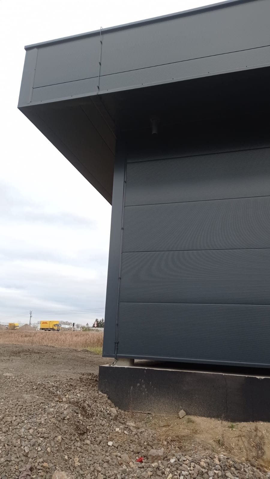 Szary budynek przemysłowy z widoczną instalacją odgromową na dachu, na tle nieba i oddalonych samochodów dostawczych.