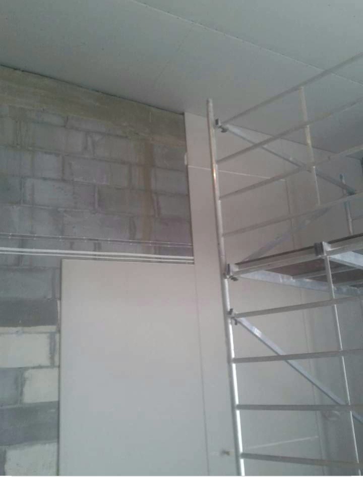 Montaż białej płyty na ścianie z pustaków, widoczne rusztowanie i sufit podwieszany w trakcie instalacji.