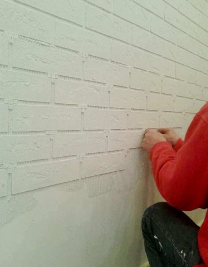 Nakładanie dekoracyjnych paneli ściennych imitujących cegłę na przygotowaną ścianę, widoczne ślady kleju i fragment osoby w czerwonej bluzie.