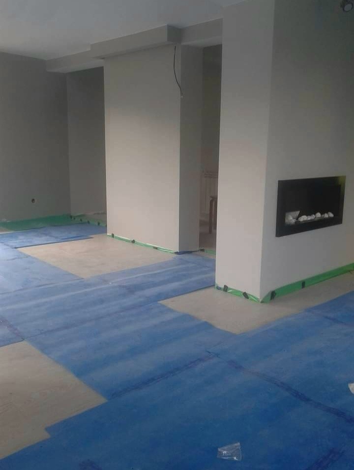 Przygotowanie podłoża pod panele: podkład izolacyjny w kolorze niebieskim rozłożony na części powierzchni w szarym pomieszczeniu z kominkiem, zabezpieczone ściany taśmą malarską.