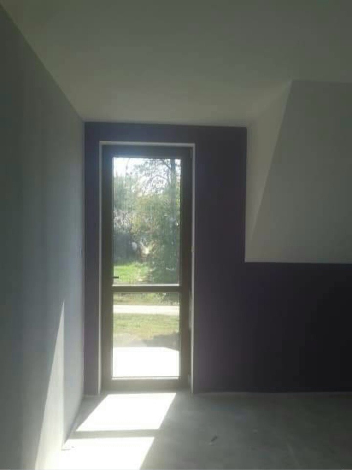 Wnętrze pokoju po malowaniu ścian na szaro i fioletowo, z zamontowanymi drzwiami balkonowymi z widokiem na ogród, jasne promienie słoneczne wpadają do środka.