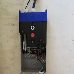 Otwarta obudowa sterownika z widocznymi przewodami, elementami elektronicznymi i przyciskiem awaryjnym na szarej ścianie.