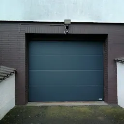 Zamknięta, segmentowa brama garażowa w kolorze antracytowym, zamontowana w brązowej, ceglanej wnęce garażu. Nad bramą widoczna lampa oświetlająca wjazd.