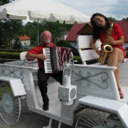 Muzyk grający na akordeonie i kobieta trzymająca saksofon siedzą na białym powozie na tle domów i zieleni.
