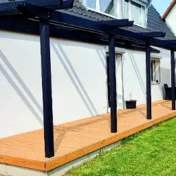 Długi, drewniany taras z czarną pergolą przylegający do białego domu, z widocznymi cieniami słupów na ścianie, mała roślina w doniczce na tarasie, trawnik obok.