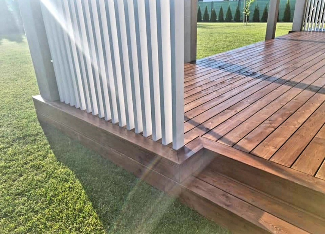 Fragment drewnianego tarasu z jasnymi, pionowymi listwami balustrady, widok z poziomu trawnika w słoneczny dzień.