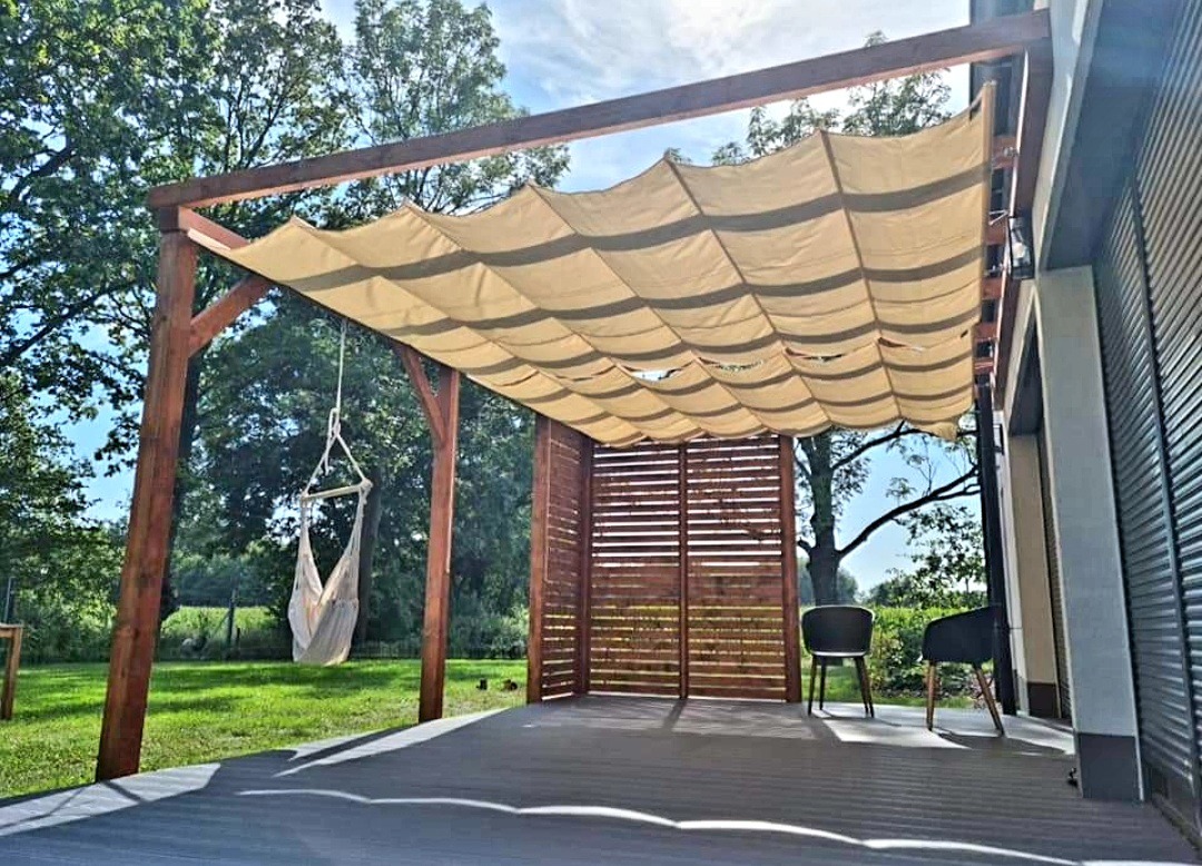 Zadaszony taras z drewnianą konstrukcją, beżową tkaniną falistą jako osłoną przeciwsłoneczną, hamakiem wiszącym z boku i drewnianą ścianką działową, na tle zielonego trawnika i drzew.