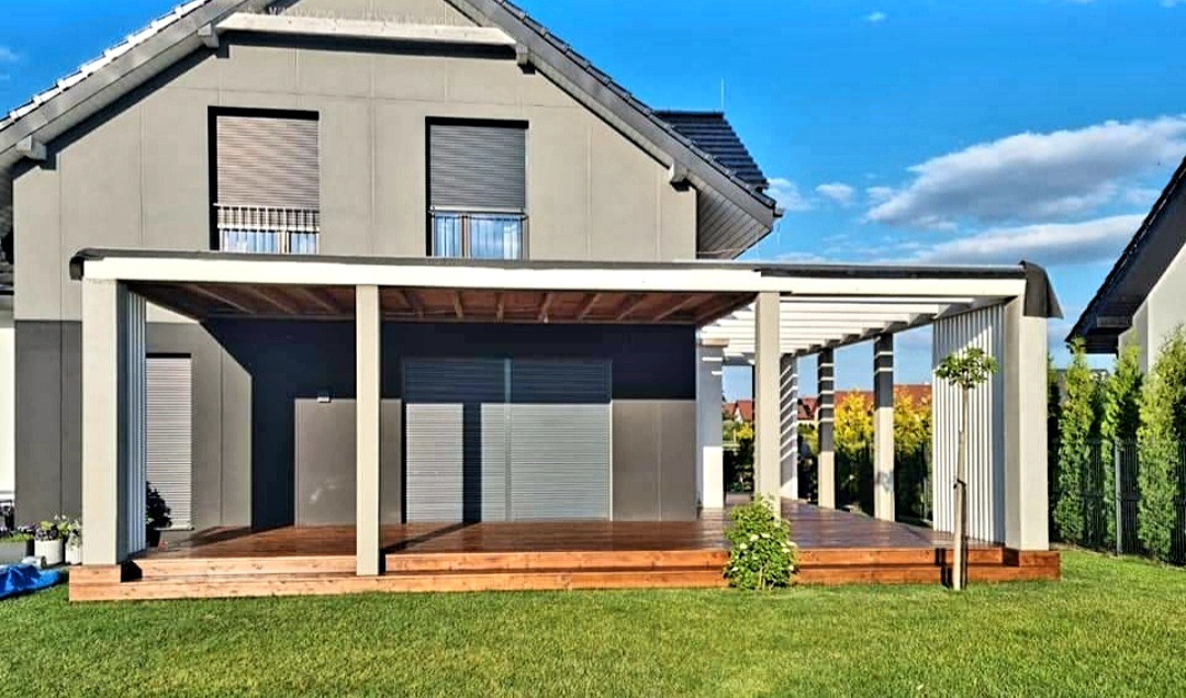 Nowoczesna pergola z drewnianym tarasem przylegająca do szarego domu z roletami zewnętrznymi, widok na zadbany trawnik i drzewa w tle.