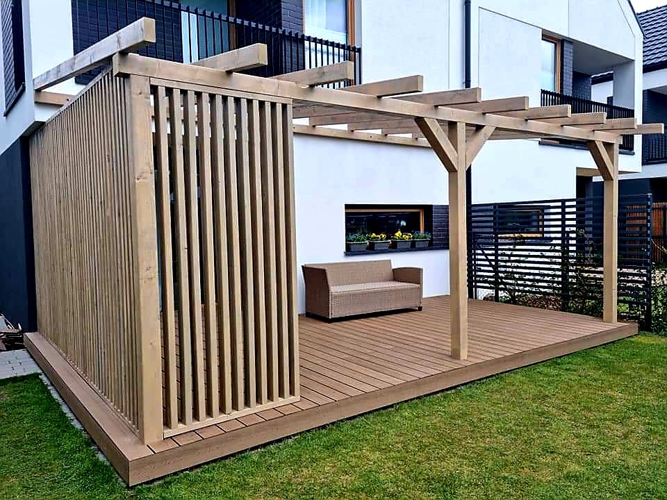 Drewniana pergola z ażurową ścianką boczną na tarasie kompozytowym, obok nowoczesnego domu z oknem z kwiatami.