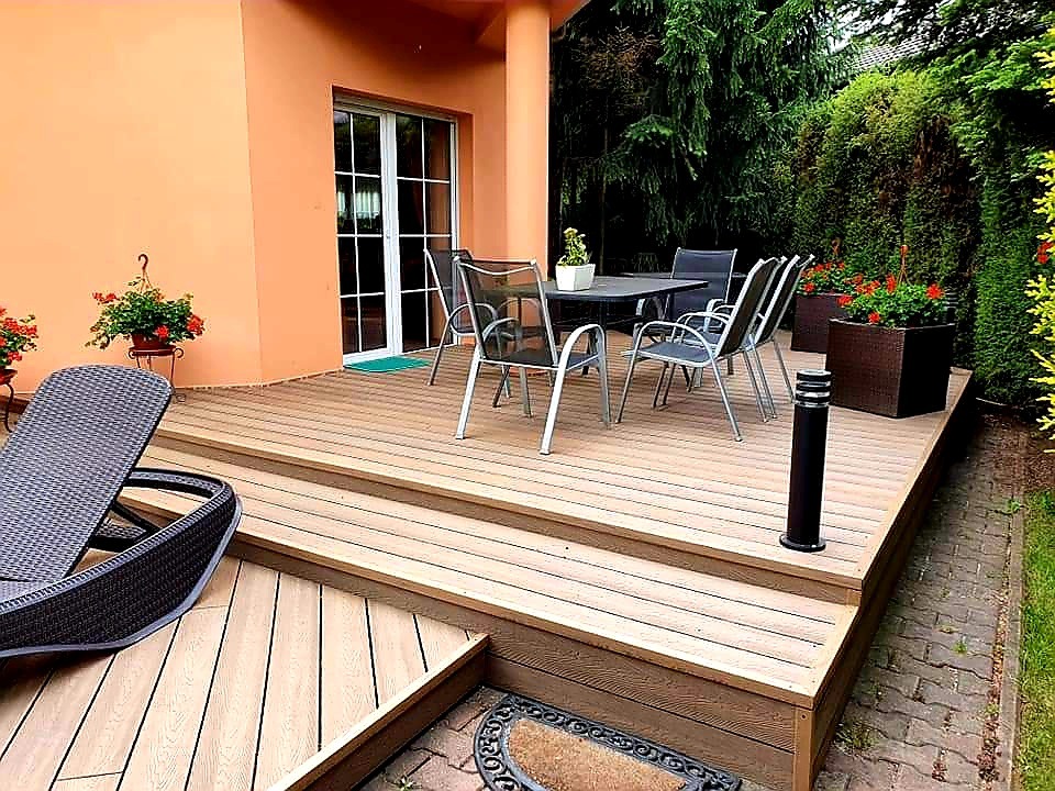 Jasny taras z desek kompozytowych z niskimi schodami, wyposażony w ciemne meble ogrodowe i lampę stojącą, otoczony zielenią.