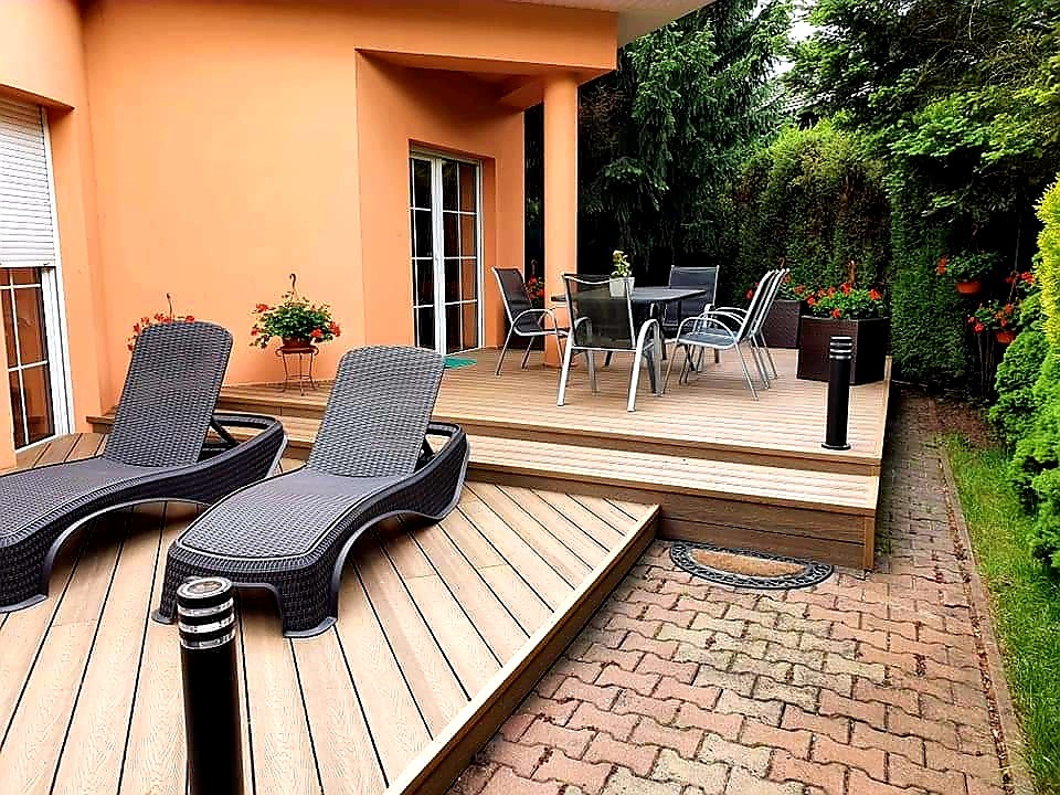 Jasny taras z desek kompozytowych z dwoma leżakami z rattanu, stolikiem z krzesłami i donicami z kwiatami, przylegający do pomarańczowego budynku z oknami i zielonego ogrodu.