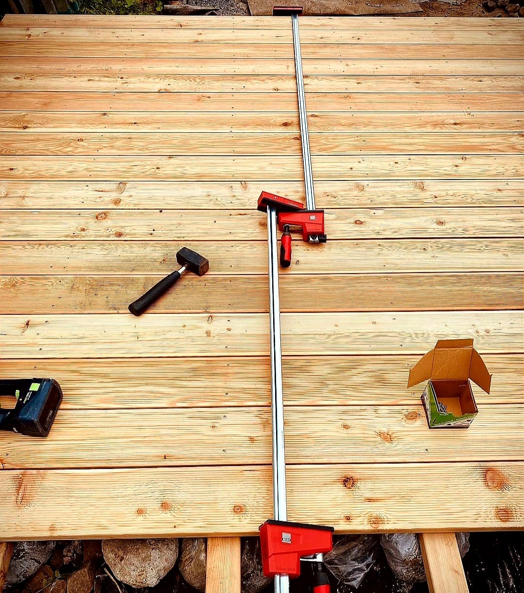 Deski tarasowe mocowane za pomocą ścisku stolarskiego, widoczny młotek i otwarte pudełko z wkrętami na tle nowo ułożonego tarasu.