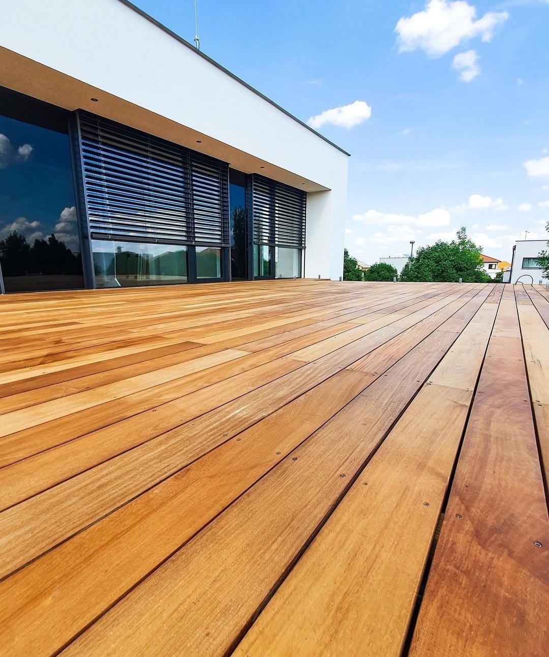 Nowoczesny taras drewniany z desek w ciepłym odcieniu brązu, przylegający do budynku z dużymi oknami z roletami zewnętrznymi, widok z niskiej perspektywy w słoneczny dzień.