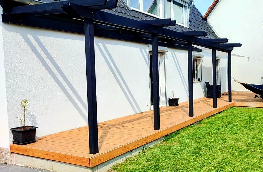Długi, drewniany taras z czarną pergolą przylegający do białego domu, z widocznymi cieniami słupów na ścianie, mała roślina w doniczce na tarasie, trawnik obok.
