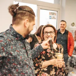 Kobieta w okularach przemawia do mikrofonu, trzymając drinka, podczas imprezy z udziałem kilku osób w pomieszczeniu.