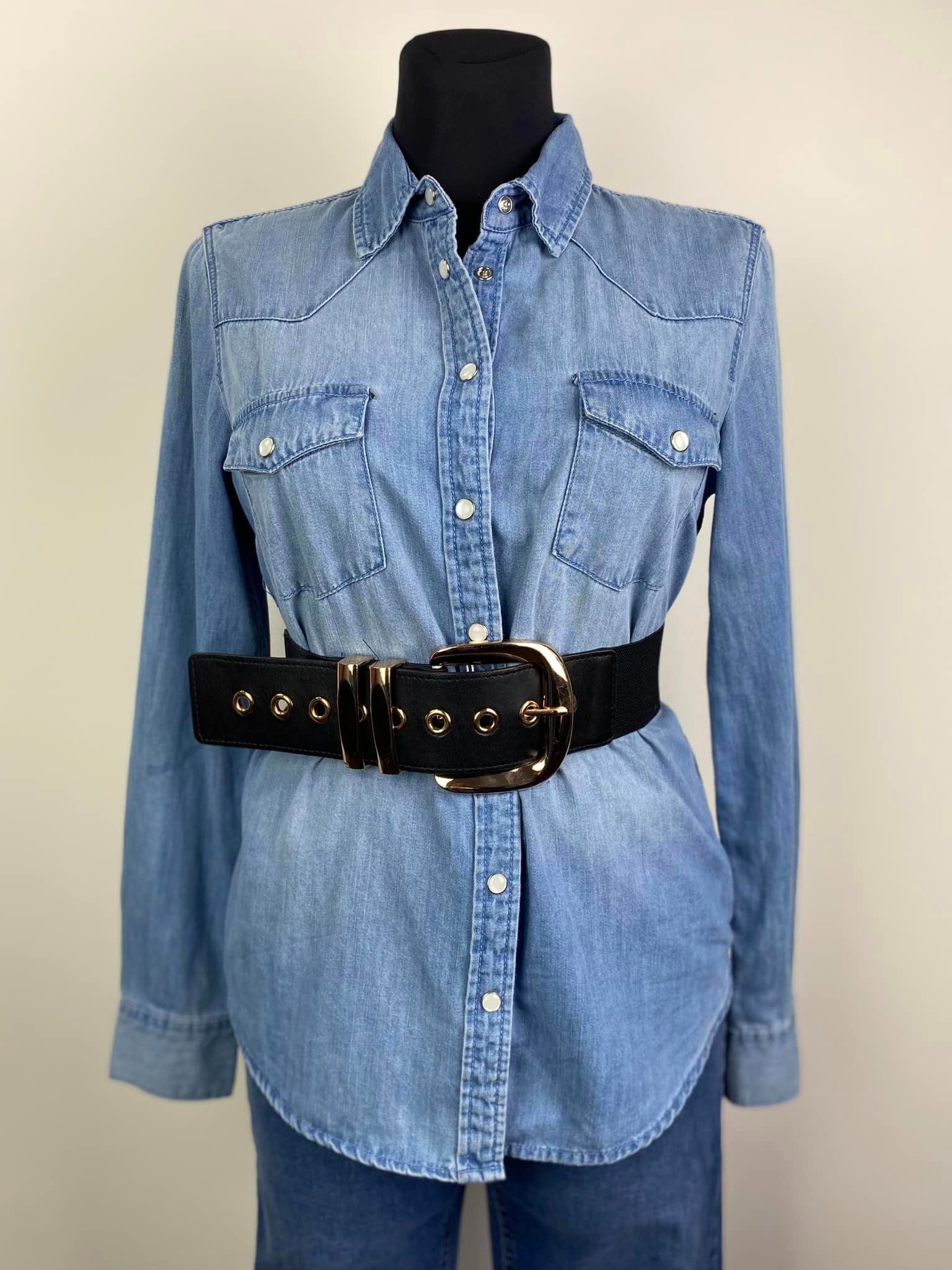 Jasna jeansowa koszula damska z długim rękawem, zapinana na zatrzaski, z czarnym paskiem z dużą złotą klamrą, prezentowana na manekinie.