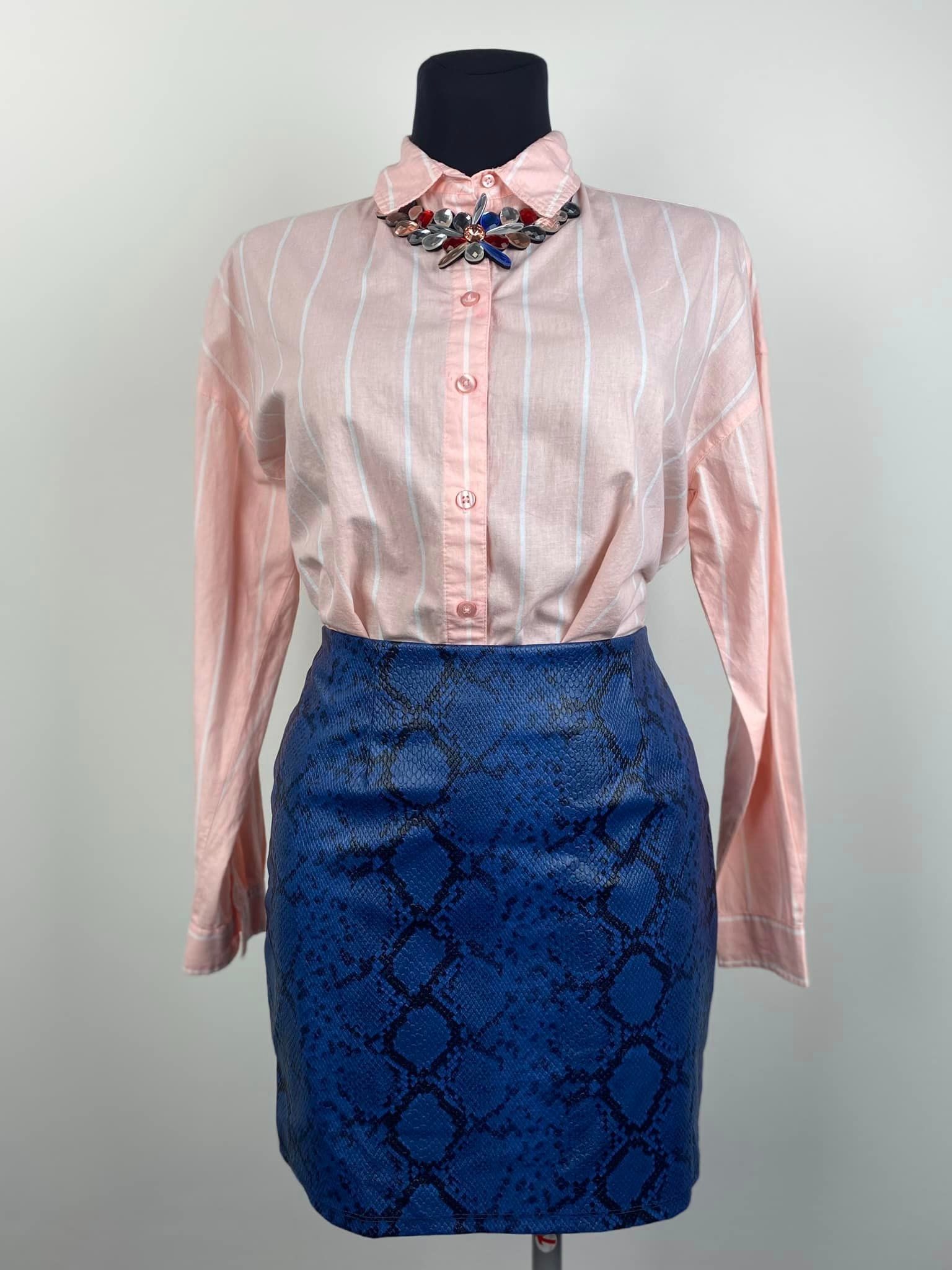 Różowa koszula w białe paski z ozdobnym broszko-kołnierzykiem i niebieska spódnica imitująca skórę węża na manekinie.