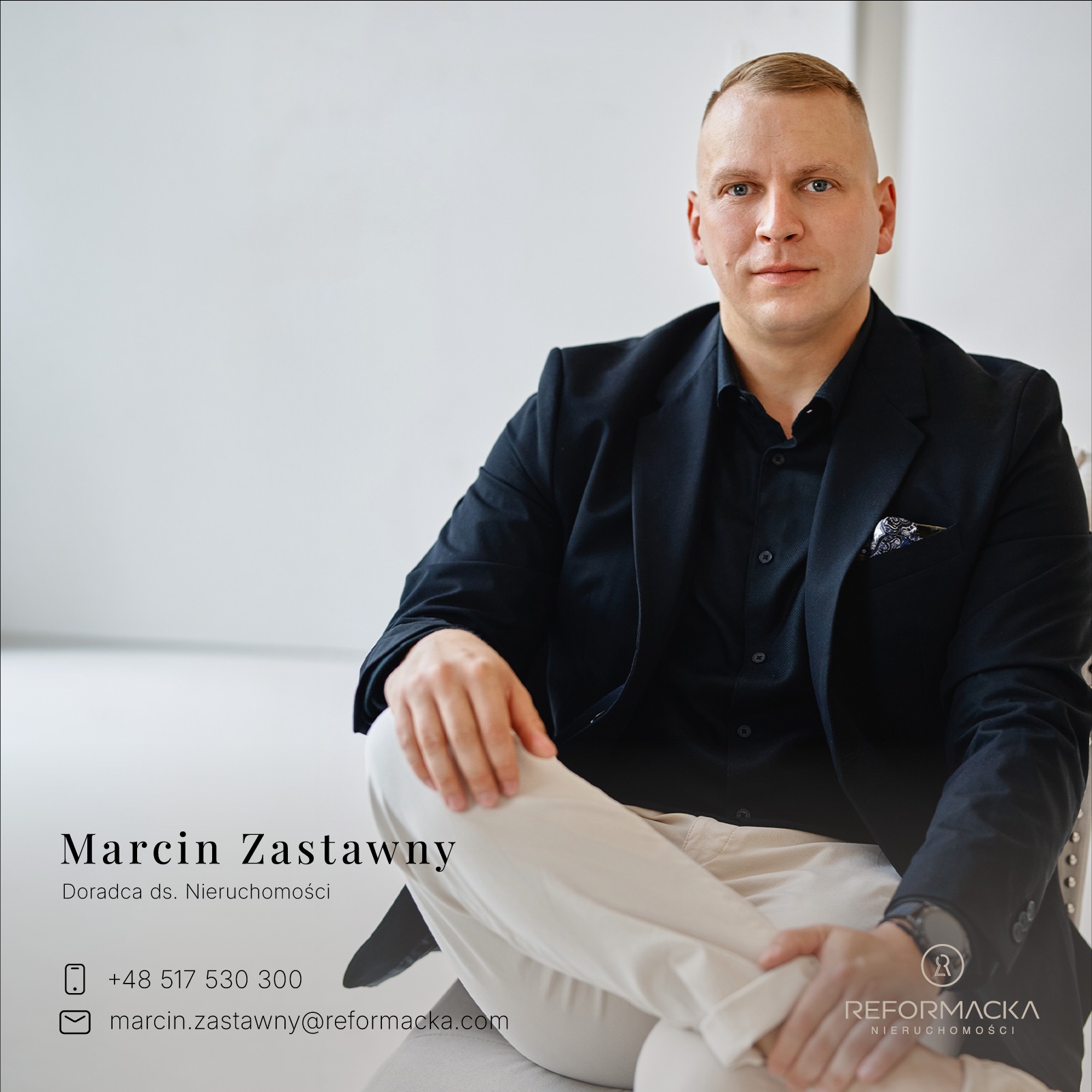 Portret mężczyzny w czarnej marynarce i beżowych spodniach, siedzącego na krześle na białym tle, z widocznym logo agencji nieruchomości w prawym dolnym rogu.