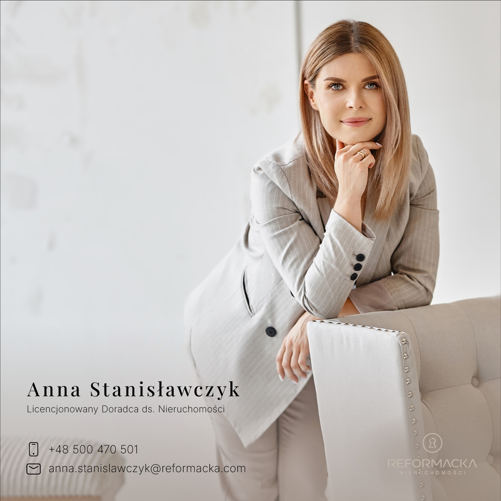 Portret kobiety w szarym garniturze, opierającej się o jasny fotel z ozdobnymi nitami, z delikatnym uśmiechem i pierścionkami na palcach.