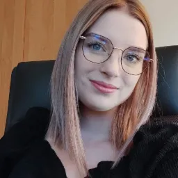 Uśmiechnięta kobieta w okularach, siedząca w biurze, z szafką i rośliną w tle.