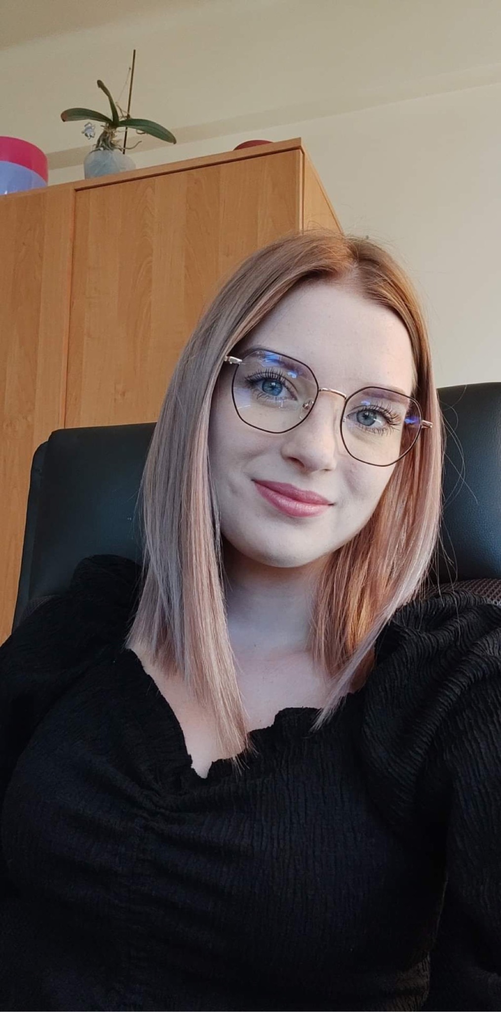 Uśmiechnięta kobieta w okularach, siedząca w biurze, z szafką i rośliną w tle.