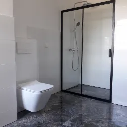 Nowoczesna łazienka z białymi płytkami, podwieszaną toaletą i kabiną prysznicową z czarną ramą na ciemnej, wzorzystej podłodze.