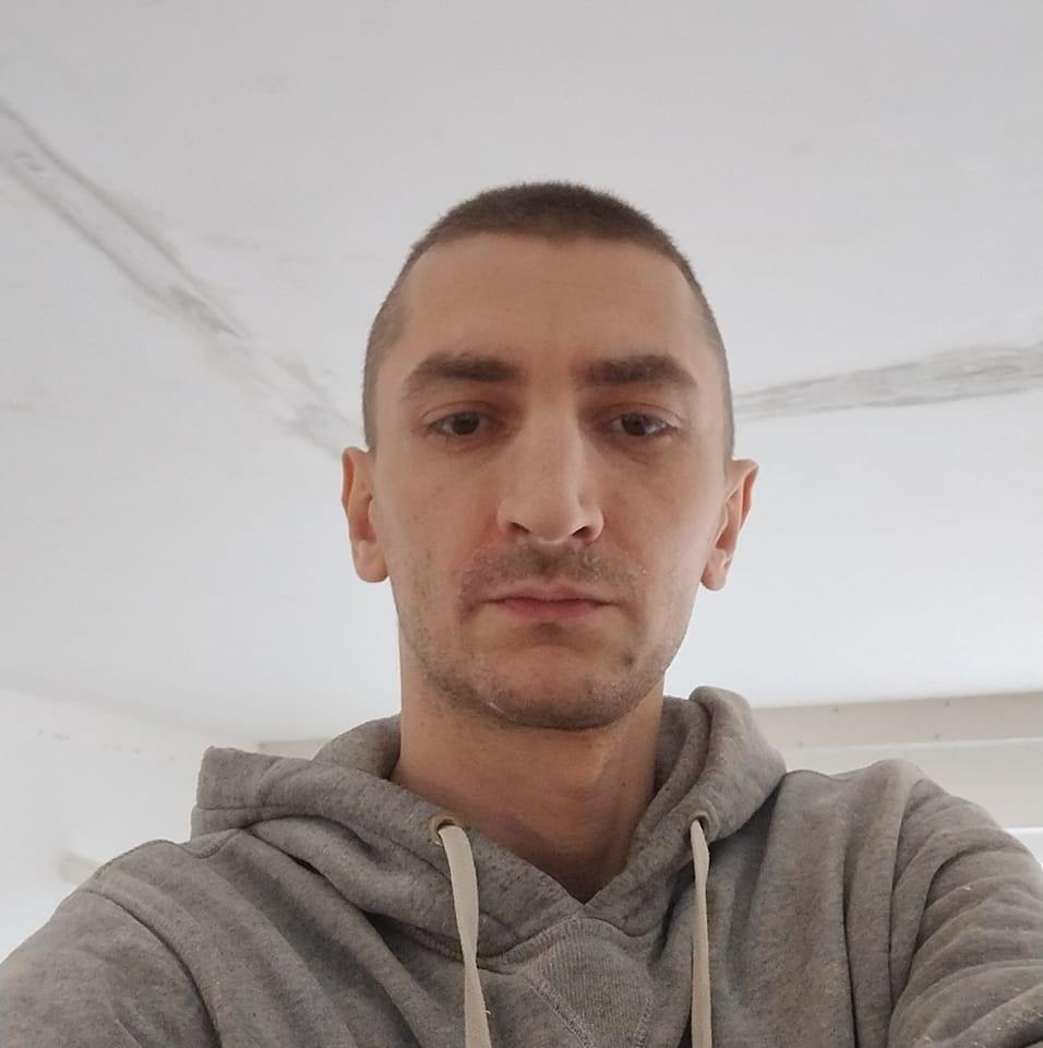 Mężczyzna w szarej bluzie z kapturem, selfie z widocznym fragmentem sufitu w trakcie szpachlowania, z widocznymi śladami po łatach.