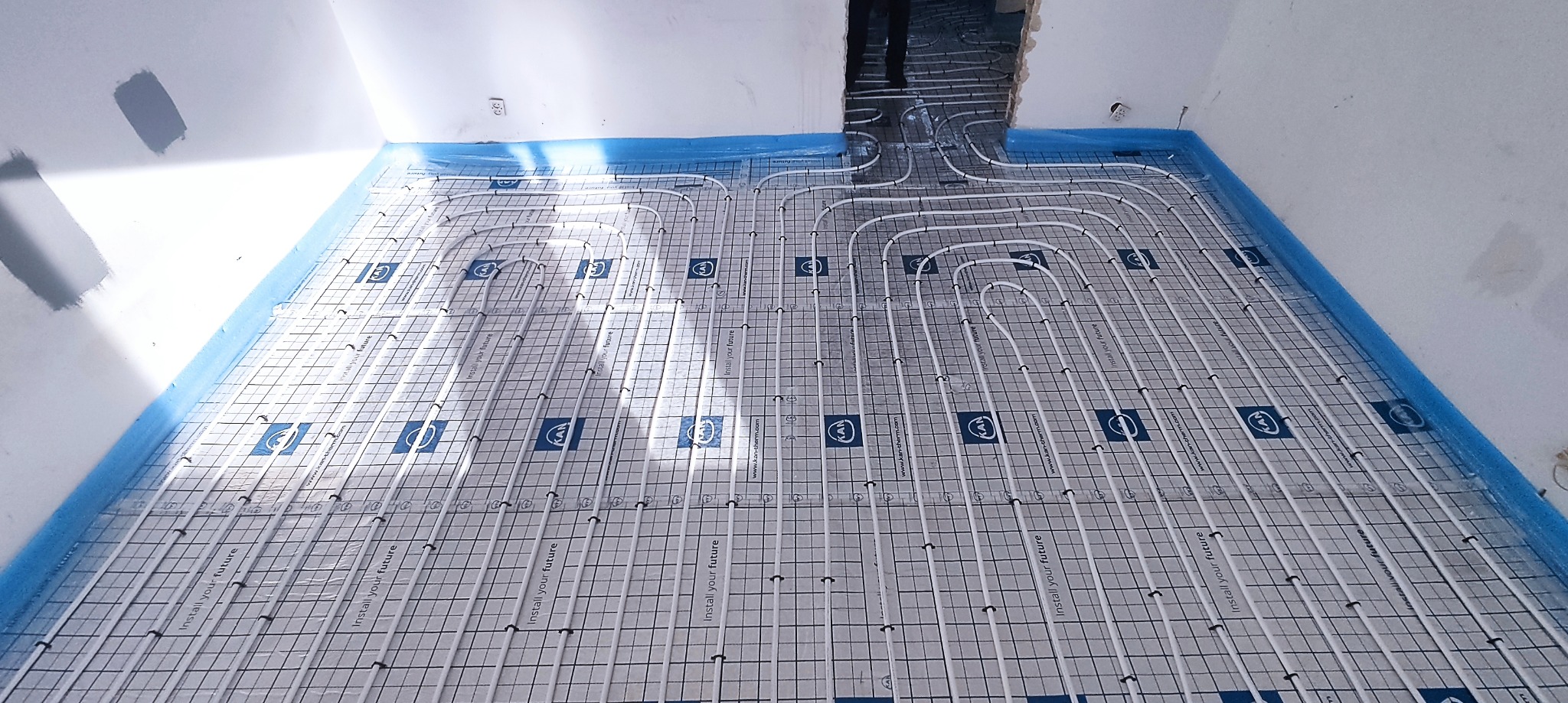 Białe rury ogrzewania podłogowego ułożone na folii z nadrukowaną siatką i logo KAN-therm, zabezpieczone niebieską folią malarską przy ścianach w surowym stanie.
