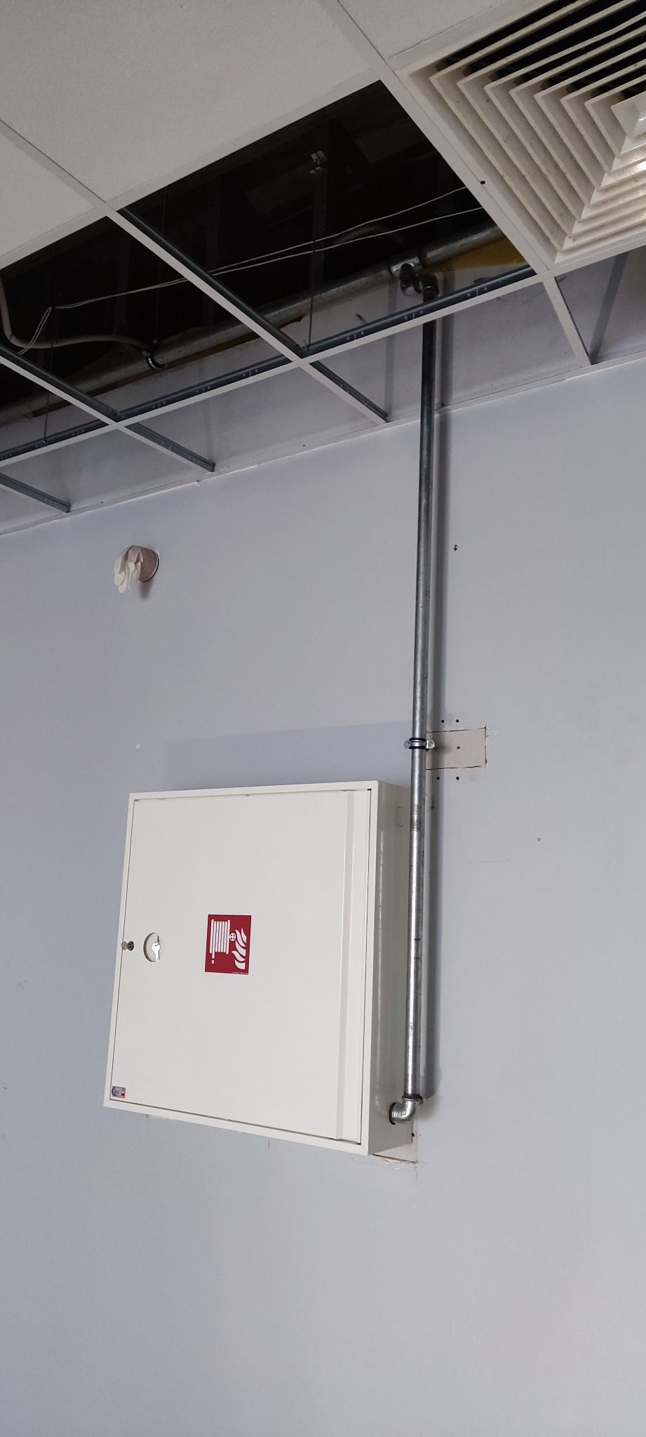 Biała szafka hydrantowa z czerwoną ikoną węża pożarniczego, zamontowana na szarej ścianie pod sufitem podwieszanym, z pionowym metalowym rurociągiem obok.