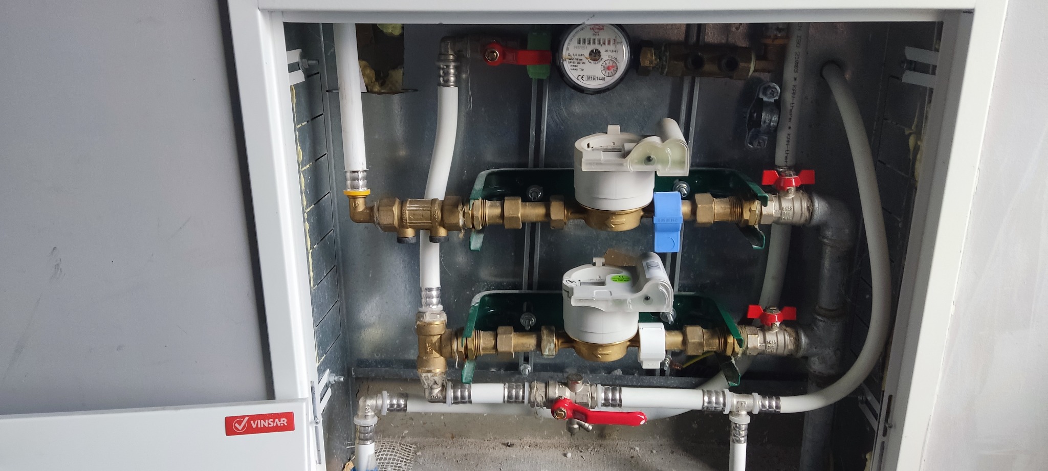 Węzeł wodomierzowy w szafce, z dwoma wodomierzami z niebieskimi i zielonymi elementami, rurami z tworzywa i mosiężnymi kształtkami, widoczne zawory kulowe z czerwonymi rączkami oraz licznik...