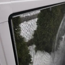 Zamontowana moskitiera ramkowa w białym oknie, widok na zieleń za oknem.