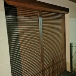 Zewnętrzna brązowa roleta z poziomych lameli, zamontowana na ścianie budynku, częściowo zasłonięta, widoczne refleksy światła.