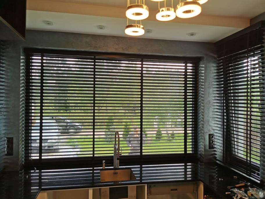 Ciemne drewniane żaluzje w nowoczesnej kuchni z widokiem na zieleń za oknem, bateria kuchenna przed oknem.