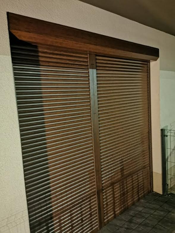 Zewnętrzna brązowa roleta z poziomych lameli, zamontowana na ścianie budynku, częściowo zasłonięta, widoczne refleksy światła.