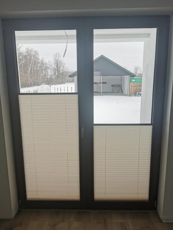 Beżowe rolety dzień-noc zamontowane w dwuskrzydłowych drzwiach balkonowych, widok na zasypane śniegiem podwórko i garaż.