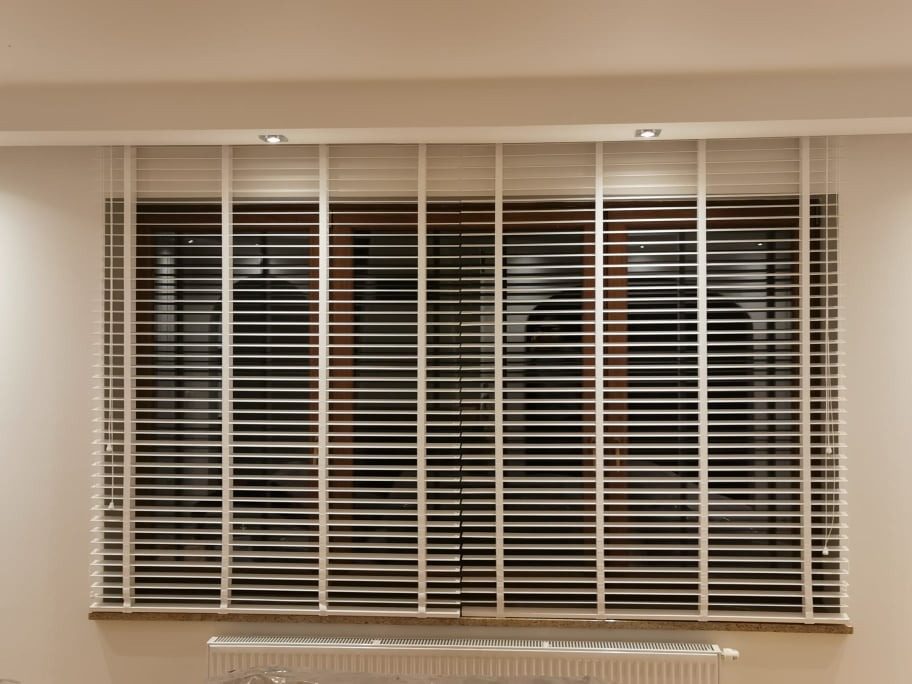 Dwa okna z białymi, poziomymi żaluzjami drewnianymi, widoczne sznurki sterujące, kaloryfer pod oknem.