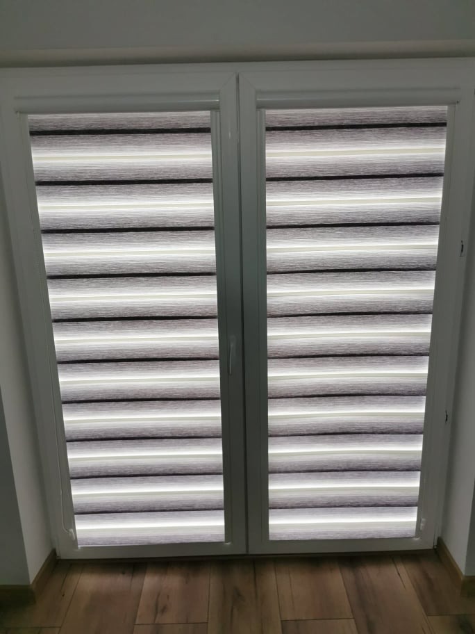 Zamknięte, dwuskrzydłowe okno z zamontowanymi roletami w odcieniach szarości i bieli, tworzącymi poziome pasy.
