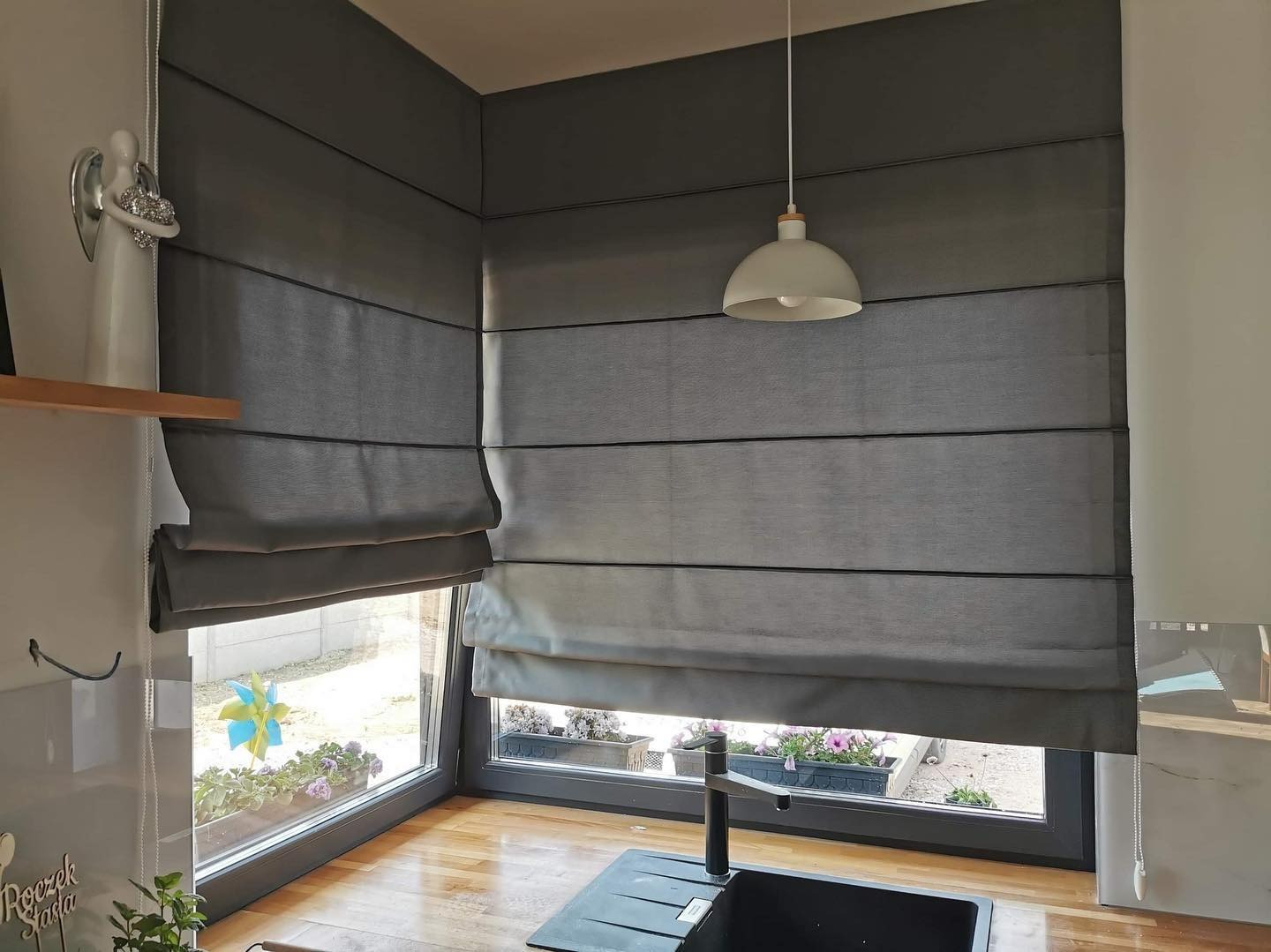 Szare, poziomo fałdowane zasłony w stylu rzymskim zamontowane w oknach nad kuchennym zlewem, widoczne kwiaty w doniczkach na parapecie.