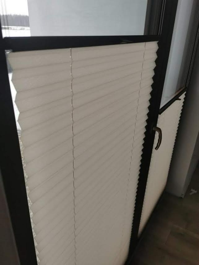 Zamknięta, plisowana osłona okienna w kolorze kremowym, zamontowana w ciemnej ramie okiennej, widoczny fragment uchwytu.