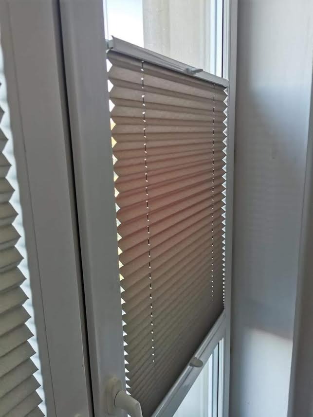 Beżowa roleta plisowana zamontowana w oknie PCV, częściowo zasłonięta, widoczne delikatne załamania materiału i mechanizm sterujący.