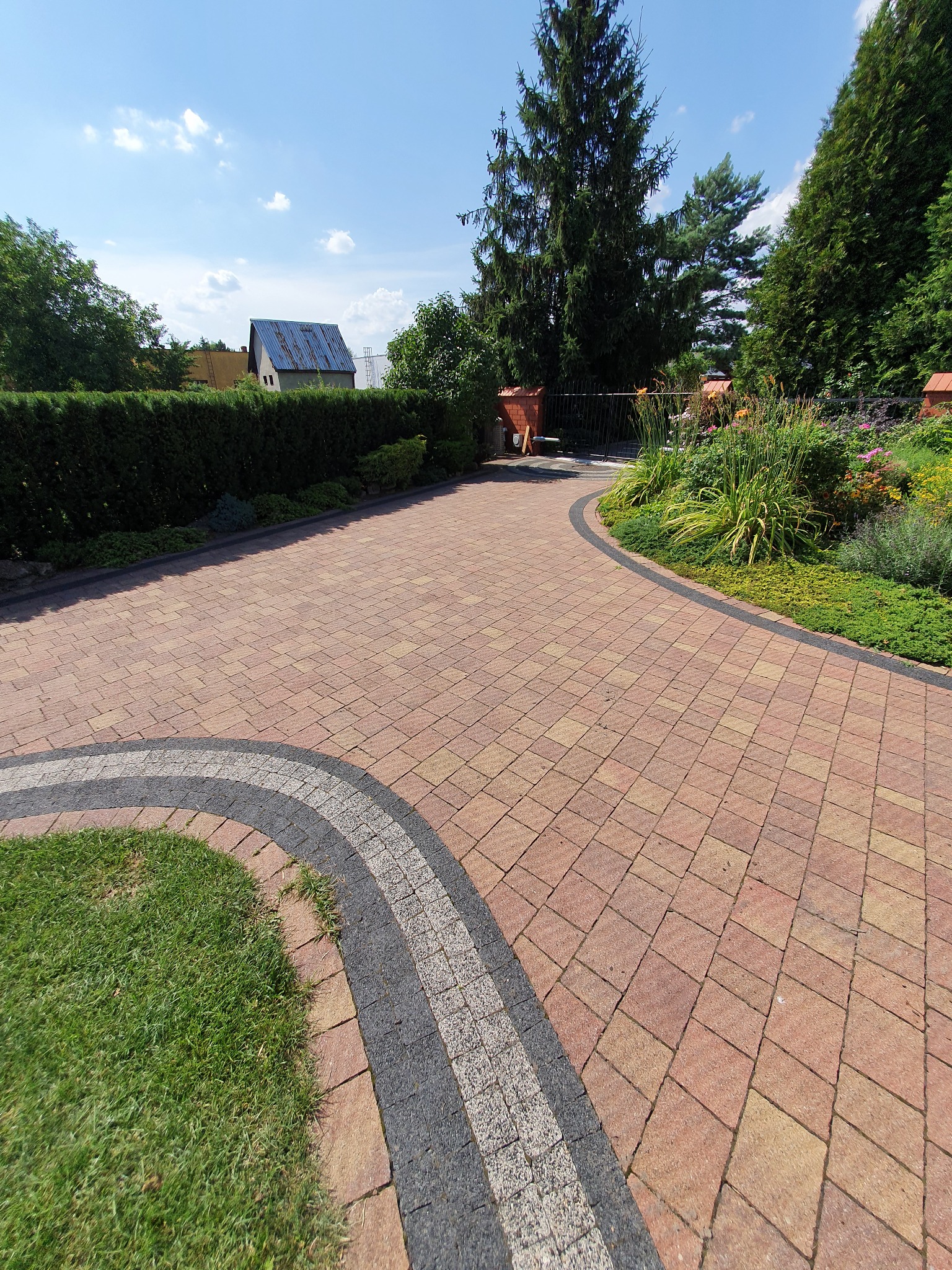 Czerwono-brązowa kostka brukowa ułożona w łukowaty wzór, obrzeża z szarej i białej kostki, trawnik, ogród kwiatowy i żywopłot w tle.