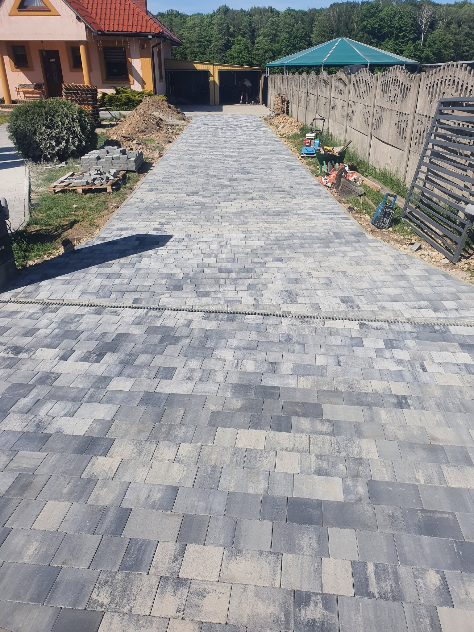 Ułożona, szara kostka brukowa na podjeździe z widocznym systemem odprowadzania wody, narzędzia budowlane i ogrodzenie w tle.