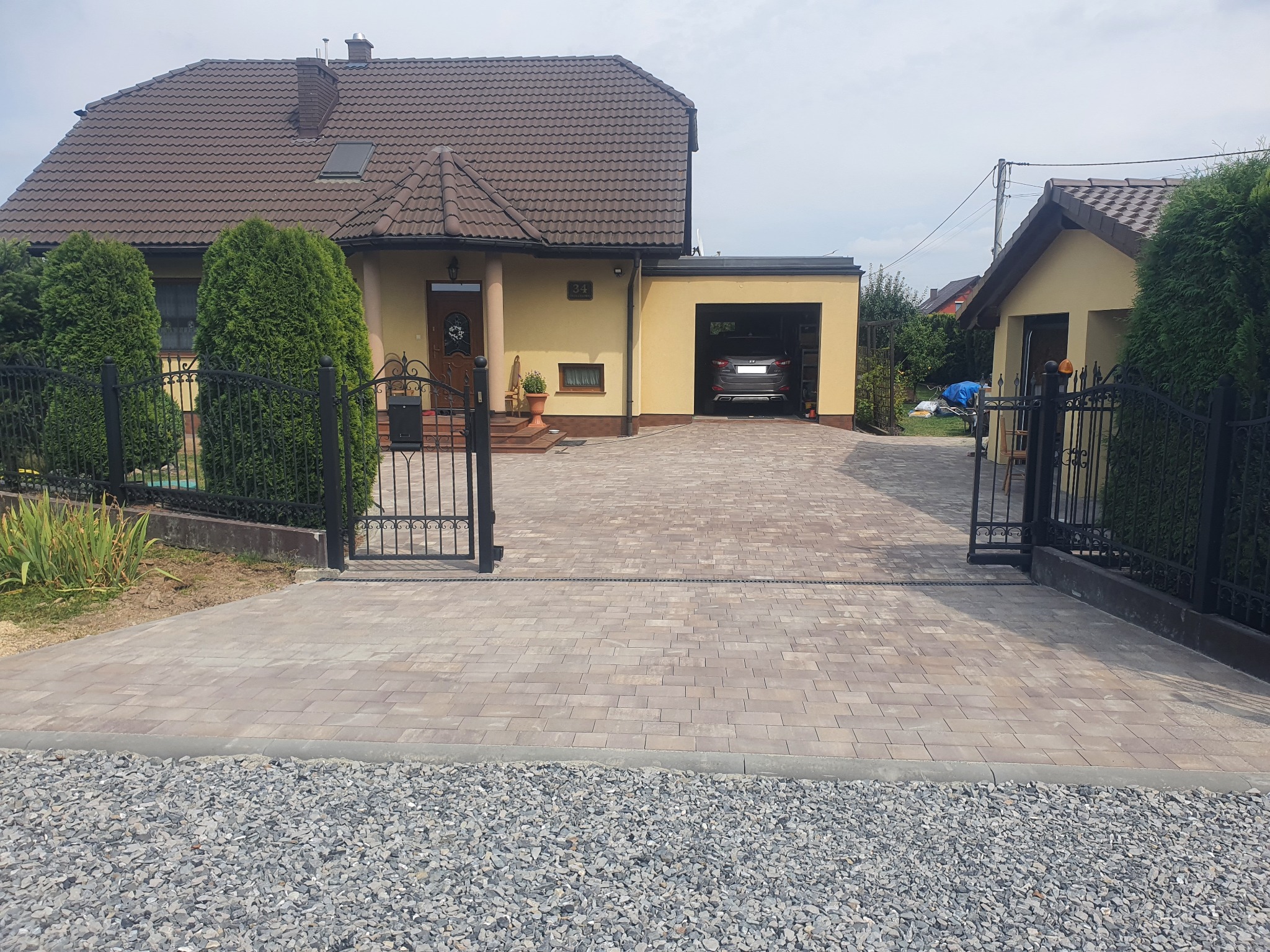 Szeroki podjazd z beżowej kostki brukowej prowadzący do domu z garażem, widoczne czarne, metalowe ogrodzenie i szara podsypka kamienna.