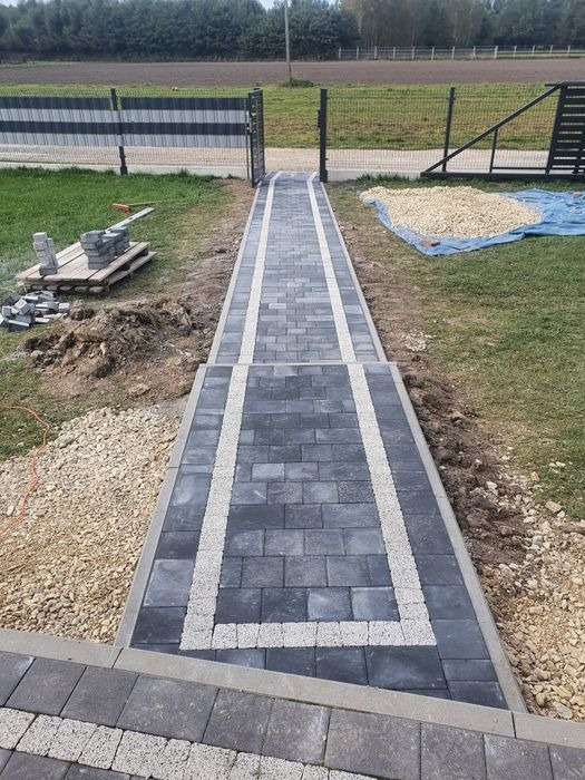 Ułożona ścieżka z szarej kostki brukowej z białymi obramowaniami prowadząca do furtki w ogrodzeniu, widoczny stos kostki na palecie i pryzma żwiru na plandece.