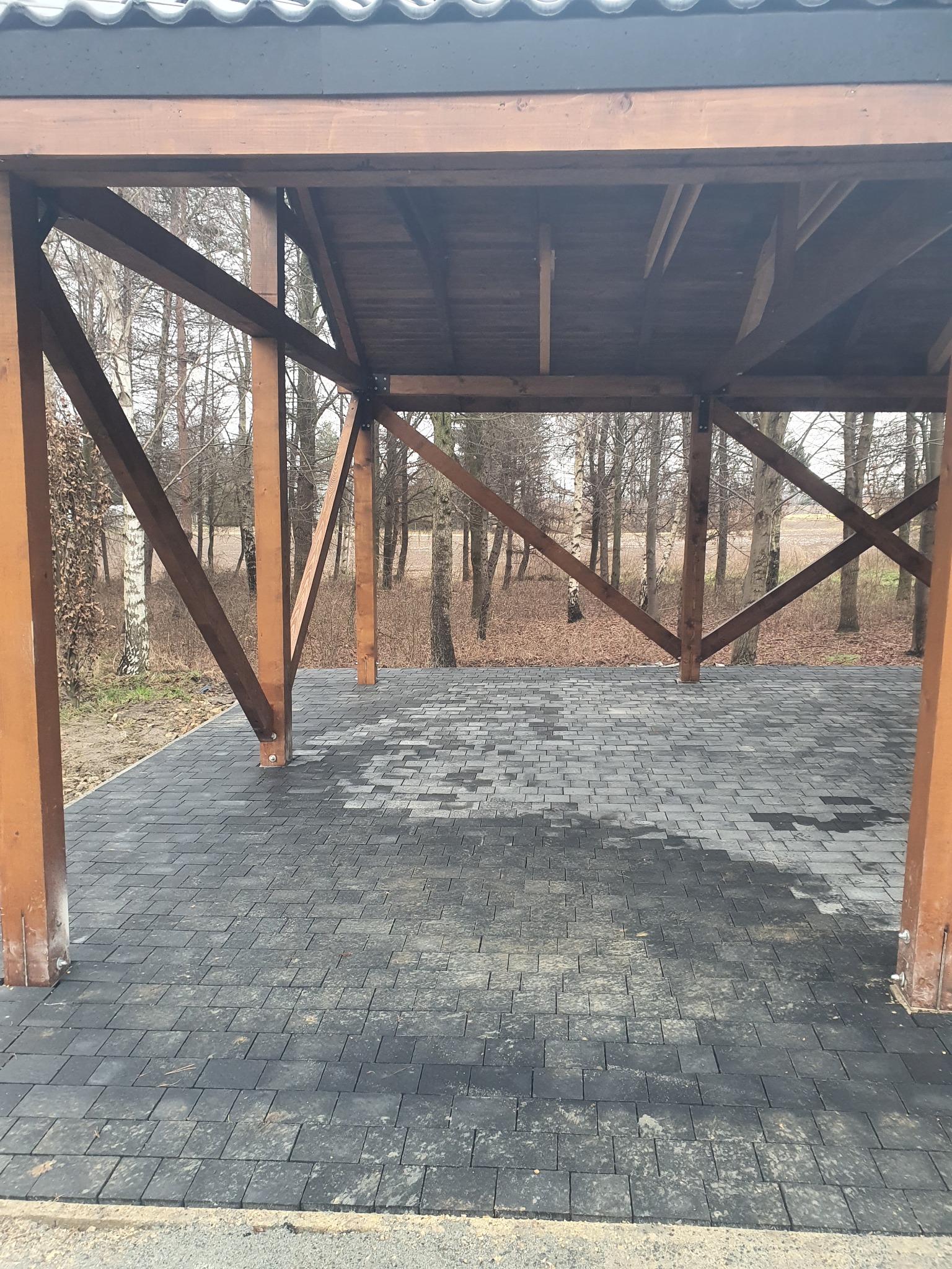 Zadaszona wiata z drewnianą konstrukcją i podłogą wyłożoną szarą kostką brukową w jodełkę, widok na drzewa w tle.
