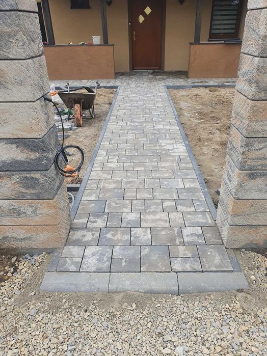 Ułożony chodnik z szarej kostki brukowej prowadzący do drzwi wejściowych domu, z widocznymi słupkami z piaskowca po bokach i taczką w tle.
