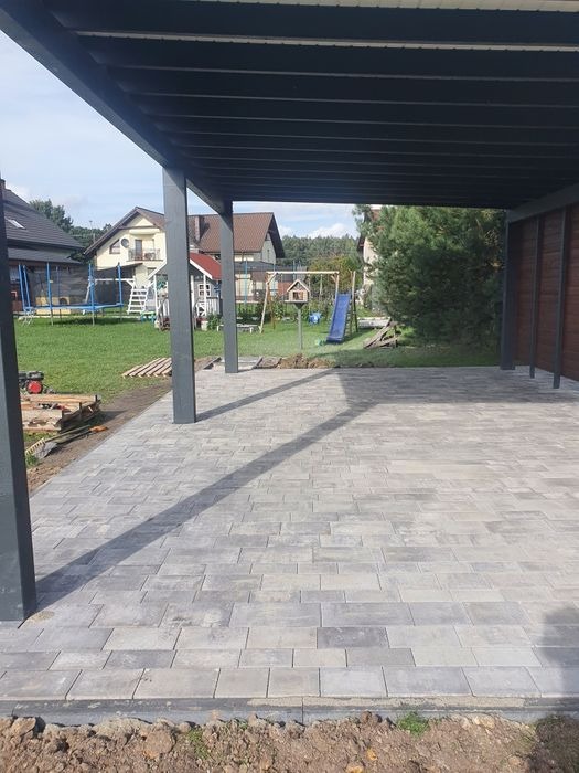 Zadaszony taras z szarej kostki brukowej ułożonej w prostokątnym wzorze, widok na ogród z placem zabaw i zabudowę jednorodzinną w tle.