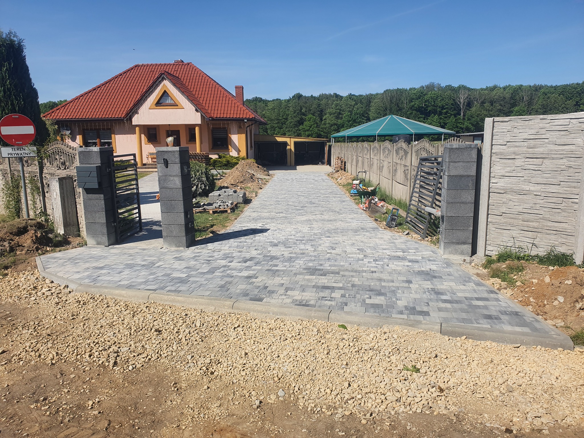 Ułożona szara kostka brukowa na podjeździe do domu jednorodzinnego z widocznym ogrodzeniem, bramą wjazdową i częściowo wykończonym terenem wokół.